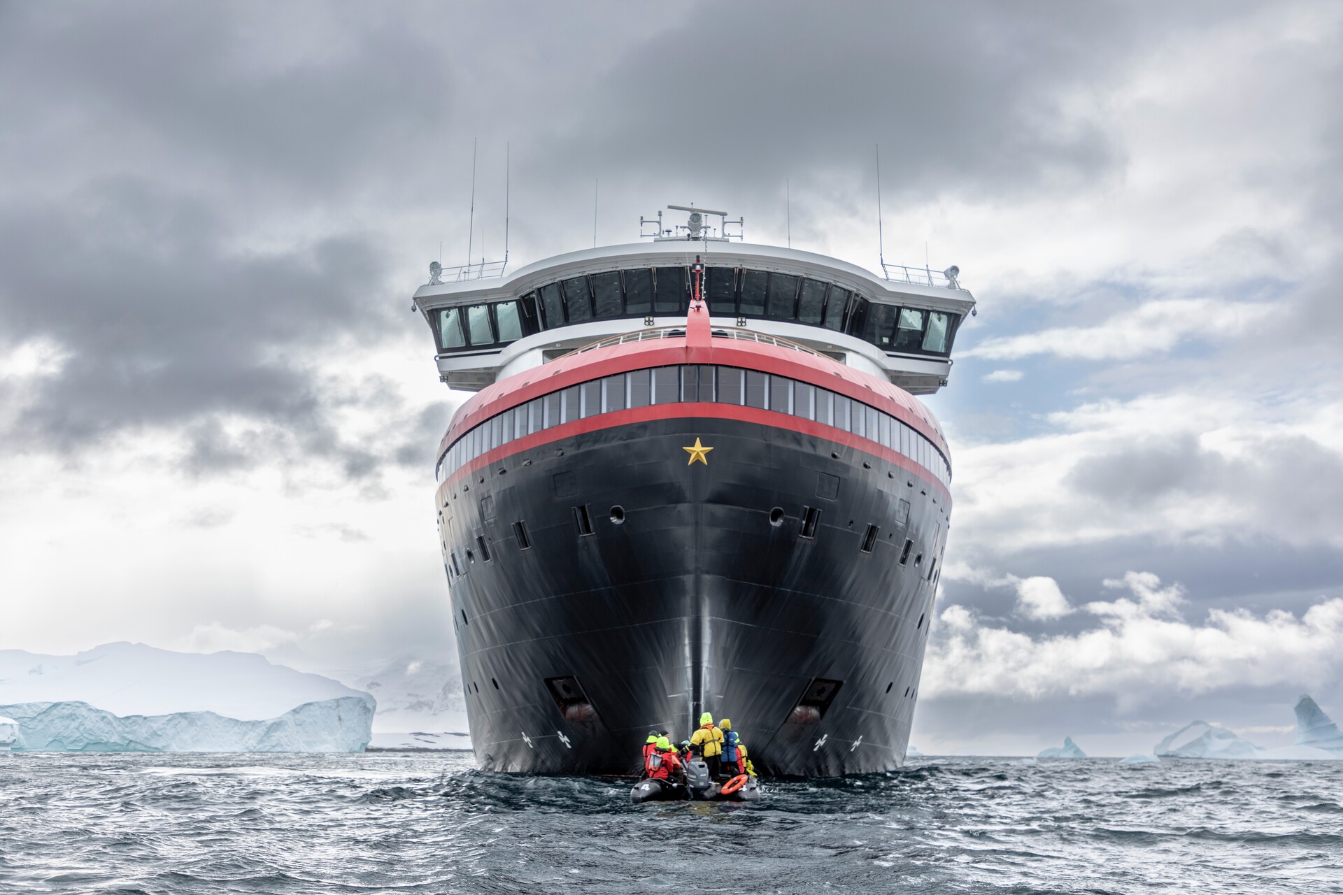 HX Expeditionsschiff von vorne gesehen, mit einem kleinen Schlauchboot und Personen im Vordergrund auf rauer See unter dramatischem, bewölktem Himmel.
