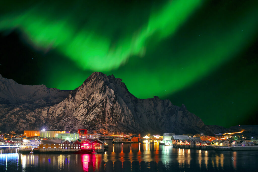 Ein nächtliches Landschaftsbild in Norwegen: Im Vordergrund spiegeln sich bunte, beleuchtete Gebäude in ruhigem Wasser. Im Hintergrund erhebt sich eine dunkle, felsige Bergkette. Am Himmel leuchten grüne Nordlichter.