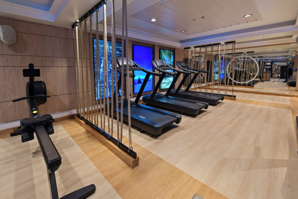 Moderner Fitnessraum der MS Roald Amundsen mit Laufbändern, die vor einer Spiegelwand aufgereiht sind und hellem Holzboden.
