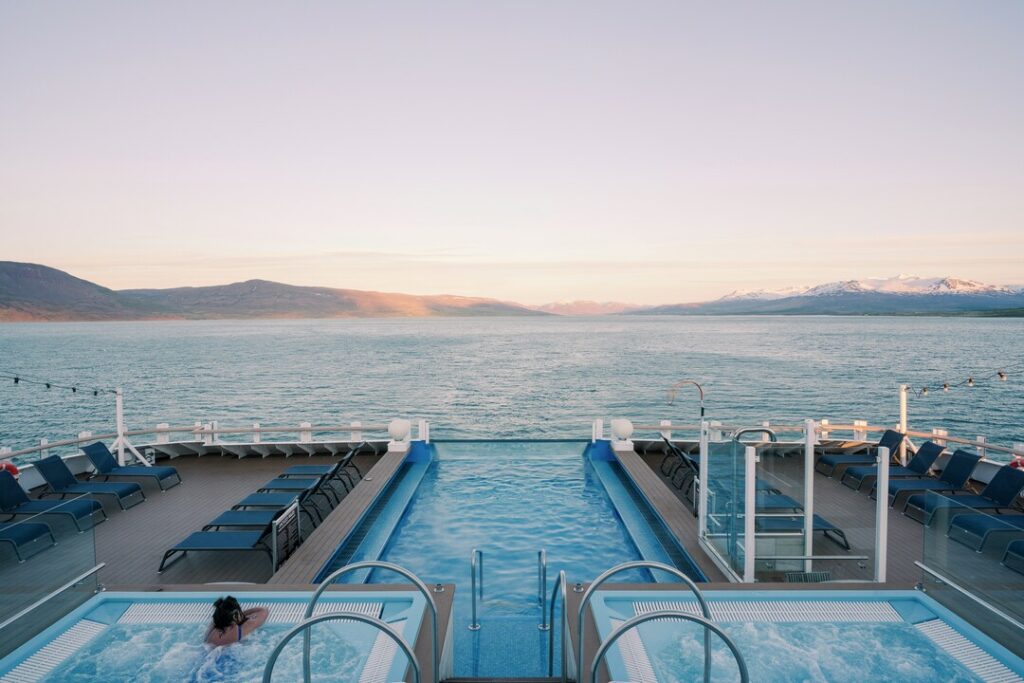 Im Vordergrund sind zwei Whirlpools und ein größerer Pool zu sehen, der direkt mit dem Heck des Schiffes endet. Im Hintergrund erstreckt sich das Meer, in der Ferne sind Berge zu sehen. Abendstimmung.