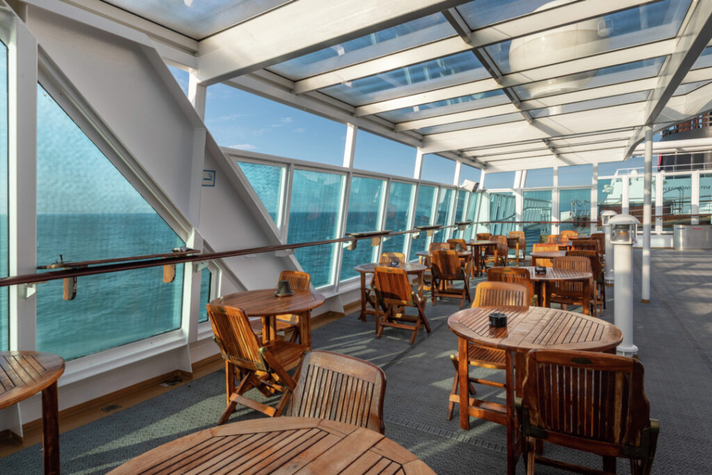 Überdachter Bereich auf der MS Maud mit großen Panoramafenstern. Die Observation Lounge ist mit Holztischen- und Stühlen ausgestattet. Das Sonnenlicht scheint durch das Glasdach und die Fenster.