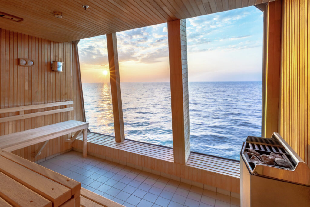Sauna auf der MS Maud mit hellem Holz verkleidet mit großen Fenstern und Blick auf das offenen Meer. Die Sonne geht am Horizont unter.
