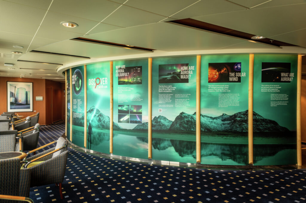 Der Raum auf dem Bild, das Science-Center, ist großflächig ausgestattet mit einer Wand, die verschiedene Informationen anzeigt und untermalt ist von einer Landschaftsgrafik mit Bergen und dem Nordlicht.