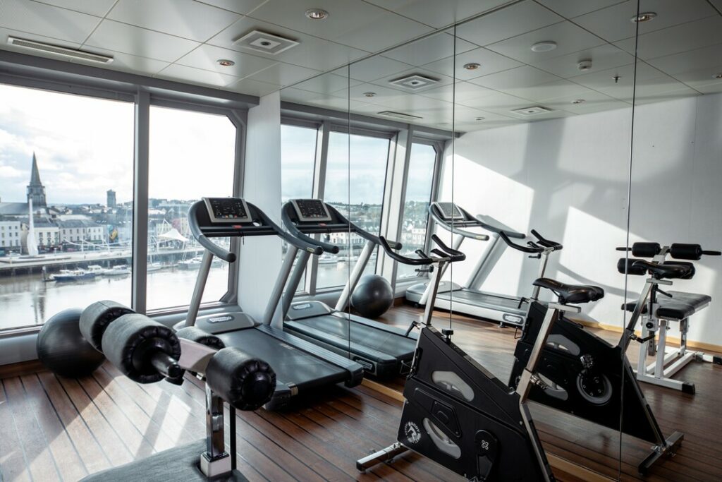 Modernes Fitnessstudio der MS Maud mit einer Reihe von Laufbändern, die vor großen Panoramafenstern aufgestellt sind. Der Blick nach draussen zeigt eine Stadt am Hafen.