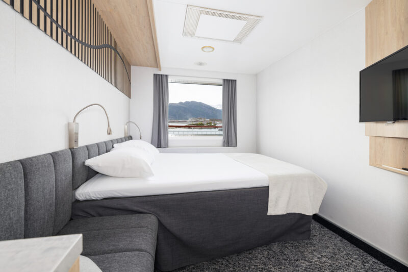 Helle Artic Außenkabine mit Doppelbett, großem Fenster mit Blick nach draußen, grauen Vorhängen und einer modernen, schlichten Einrichtung. An der Wand befindet sich ein Flachbildfernseher, die Gestaltung kombiniert warme Holzelemente mit neutralen Farben.