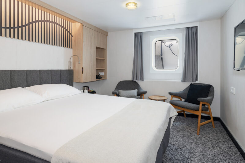 Eine modern eingerichtete Arctic Superior Kabine mit einem großen Doppelbett. Die Kabine verfügt über ein Fenster mit Blick nach draußen, das von grauen Vorhängen eingerahmt ist. Links neben dem Bett befindet sich eine Holzwand mit dekorativen Elementen sowie ein kleines Regal. Im Raum stehen zwei bequeme Sessel mit einem runden Holztisch in der Mitte. Der Boden ist mit einem grauen Teppich ausgelegt, und die Wände sind in neutralen Farben gehalten.