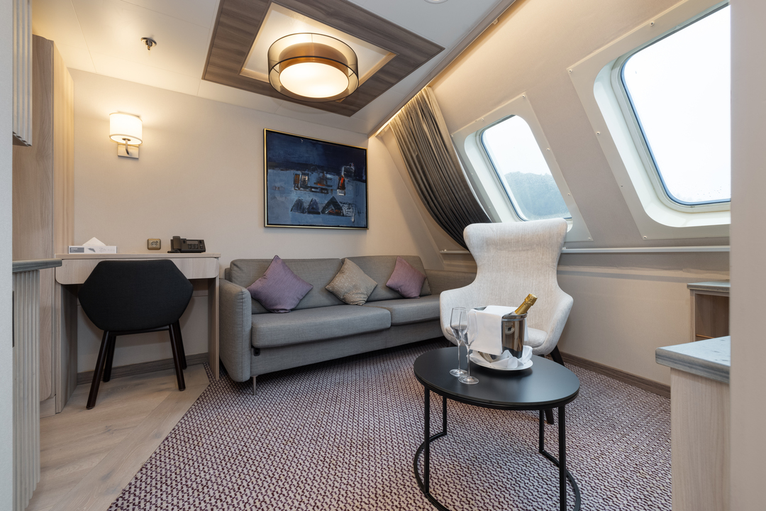 Suite der MS Otto Sverdrup mit moderner Einrichtung: Heller Raum mit einem Sofa, dekorativen Kissen, einem Sessel, einem kleinen runden Tisch mit Champagner und Gläsern, einem Schreibtisch mit Stuhl, künstlerischem Wandbild und großen Fenstern mit Blick nach draußen.