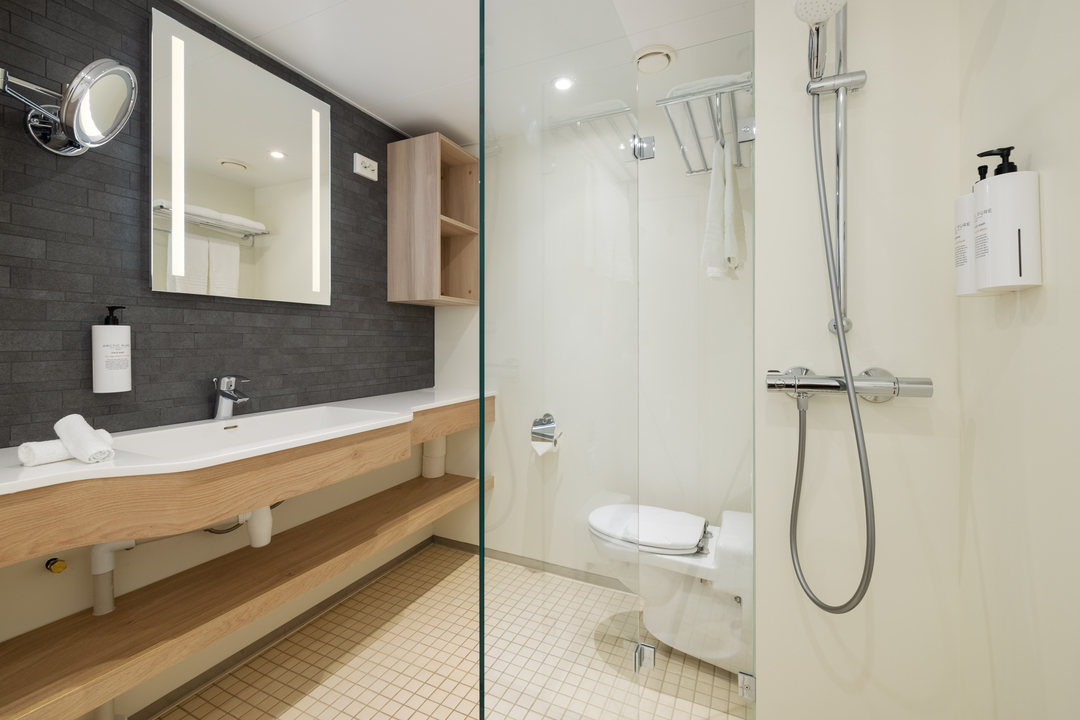 Modernes Badezimmer mit großem Waschtisch aus Holz, beleuchtetem Spiegel, dunkler Wandverkleidung, einer Dusche mit Glasabtrennung, Toilette und wandmontierten Seifenspendern.