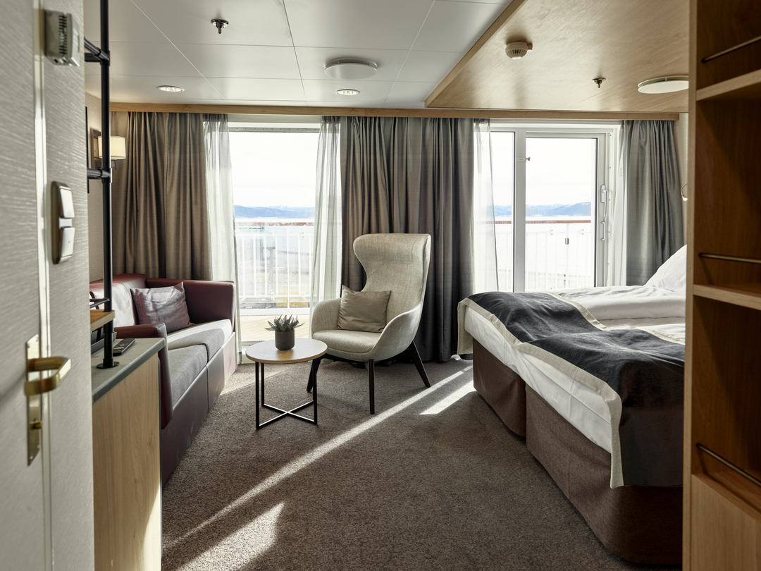 Moderne Suite der MS Otto Sverdrup mit einem Doppelbett, einem Sofa, einem Sessel, einem kleinen runden Tisch, dekorativen Kissen und großen Fenstern mit Zugang zu einem privaten Balkon.