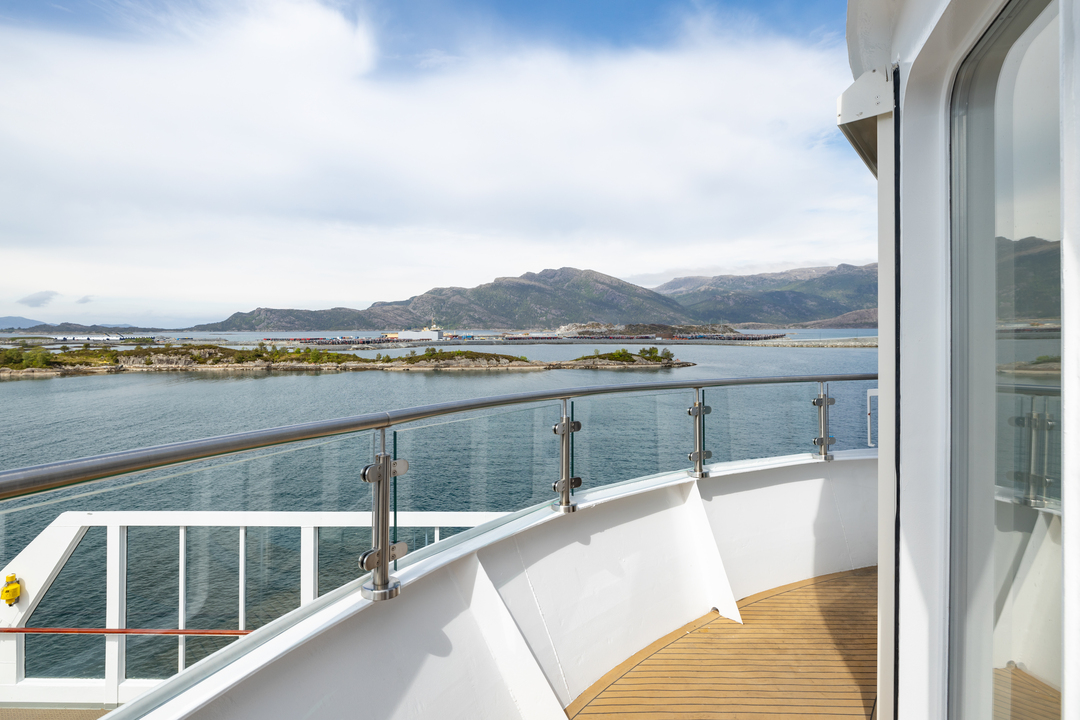 Blick vom privaten Balkon einer Grand Suite auf der MS Otto Sverdrup. Der Balkon bietet eine großzügige Aussicht auf die Landschaft mit ruhigem Wasser, Hügeln und einem strahlend blauen Himmel. Die Glasbrüstung sorgt für einen ungestörten Panoramablick.