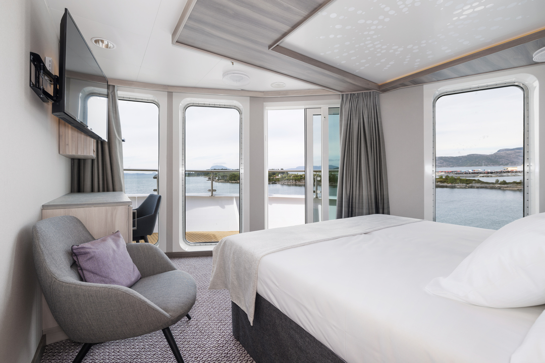 Innenansicht der Grand Suite MG auf der MS Otto Sverdrup mit einem großen Bett, gemütlicher Sitzecke und bodentiefen Fenstern. Die Suite ist modern eingerichtet, mit einem flachen Fernseher und hellen, natürlichen Farben für eine entspannte Atmosphäre.