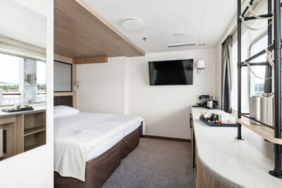 Mini Suite mit Doppelbett, Wand-TV, großzügigen Ablageflächen, einer Kaffeemaschine und einem großen Fenster für Tageslicht und Ausblick; links ein Spiegel, der den Raum optisch vergrößert.