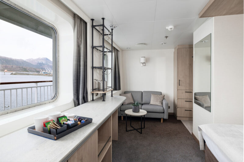 Mini Suite mit modernem Design: Großes Fenster mit Blick aufs Wasser, Sofa mit kleinem Beistelltisch, praktische Ablageflächen mit Getränken und Snacks sowie eine gemütliche Einrichtung mit Schrank und Spiegel.