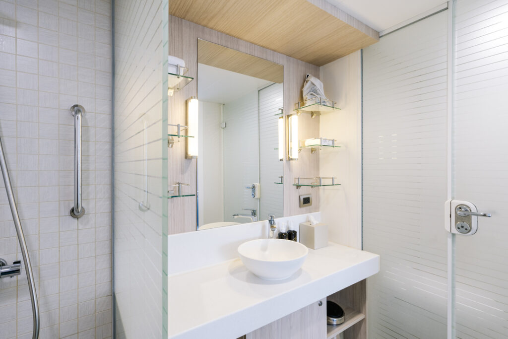 Modernes Badezimmer in der Darwin Suite auf der MS Santa Cruz II: Helle Ausstattung mit einer weißen Waschtischkonsole, einem runden Aufsatzwaschbecken, einem großen Spiegel und Glasregalen. Die Dusche verfügt über eine Handbrause und einen Haltegriff.