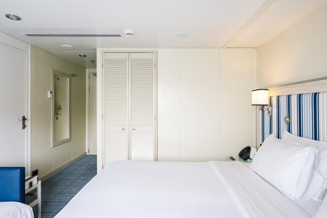 Geräumige Kabine auf der MS Santa Cruz II mit hellem, neutralem Interieur. Ein großes Bett mit weißer Bettwäsche und einem blau-weiß gestreiften Kopfteil steht im Vordergrund. An der Wand neben dem Bett befindet sich eine kleine Lampe. Im Hintergrund sieht man eine geschlossene Schranktür mit Lamellen sowie eine Zimmertür und einen Spiegel an der Wand. Der Boden ist mit einem blauen Teppich ausgelegt.