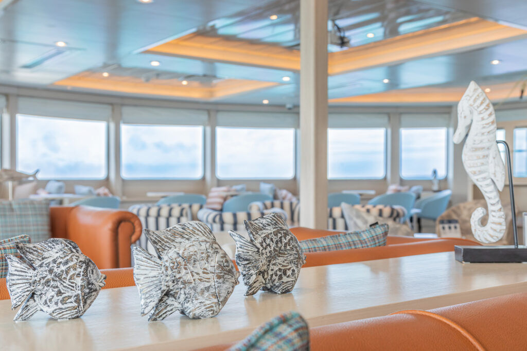 Das Bild zeigt die Expedition Lounge der MS Santa Cruz II mit großen Fenstern, die eine Aussicht auf den Himmel und das Meer bieten. Im Vordergrund stehen dekorative Figuren, darunter ein großes Seepferdchen, während im Hintergrund bequeme Sitzgelegenheiten in warmen Farben und eine moderne Beleuchtung zu sehen sind.