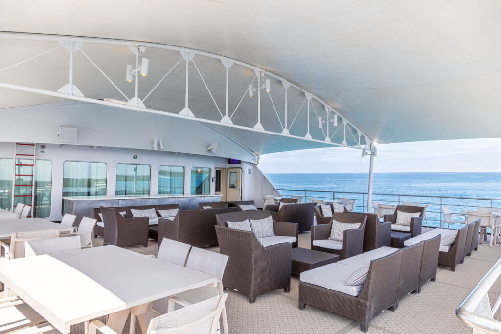 Das Bild zeigt den Observation Sky Deck der MS Santa Cruz II, ausgestattet mit gemütlichen Lounge-Möbeln aus Rattan und weißen Polstern. Unter einem schattenspendenden Dach bietet die Fläche eine entspannte Atmosphäre mit Blick auf das Meer. Im Hintergrund ist eine Bar zu sehen.
