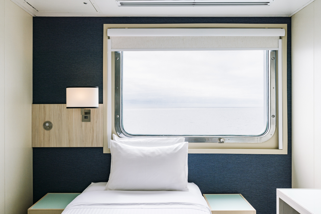 Das Bild zeigt eine Einzelkabine der MS Santa Cruz II mit modernem, schlichtem Design. Die Kabine verfügt über ein Einzelbett mit weißer Bettwäsche, eine dunkelblaue Wand, eine integrierte Lampe, einen Nachttisch und ein großes Fenster mit Blick auf das Meer.