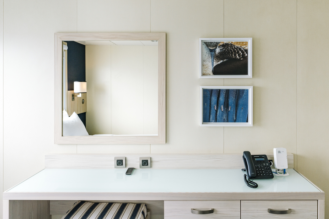 Das Bild zeigt einen Schreibtisch in einer Einzelkabine der MS Santa Cruz II mit einer Glasoberfläche und Schubladen darunter. Ein Spiegel an der Wand reflektiert einen Teil des Bettes und der Wandlampe. Darüber hängen zwei kleine Bilder mit maritimen Motiven. Auf dem Schreibtisch befinden sich ein Telefon, ein Ladegerät und eine Fernbedienung.
