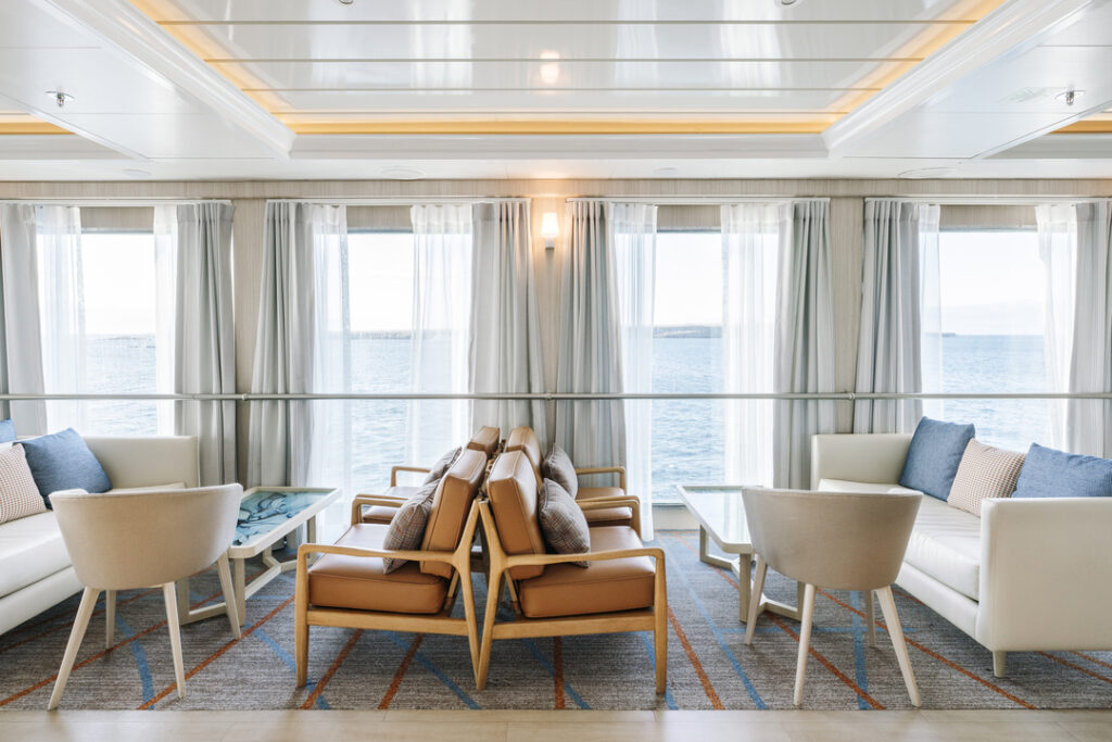 Die Panorama Lounge der MS Santa Cruz II mit großen Fenstern, die viel Tageslicht einlassen und eine Aussicht auf das Meer bieten. Es gibt mehrere Sitzgruppen mit bequemen Sesseln und Sofas in neutralen Farben, ergänzt durch Kissen in Blau- und Beigetönen. Mit hellen Vorhängen und einer dezenten Beleuchtung wirkt die Einrichtung modern.