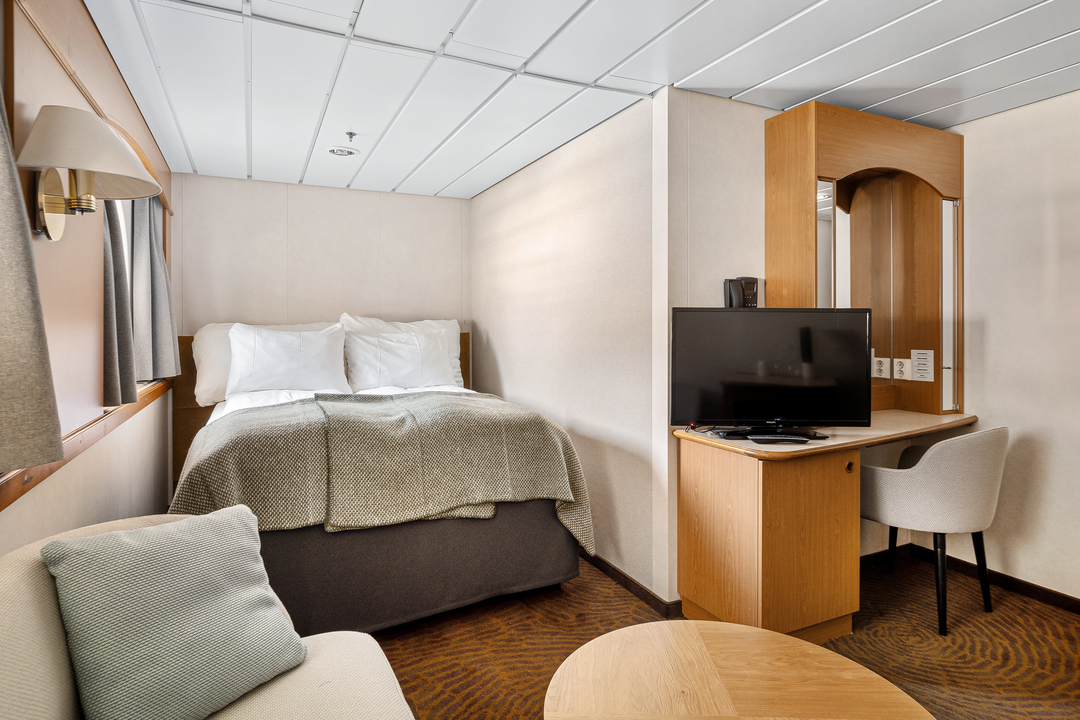 Kabine mit Doppelbett, einem Sofa, einem kleinen Tisch, einem Schreibtisch mit Stuhl und einem Flachbildfernseher. Die Einrichtung wirkt modern und gemütlich mit warmen Holztönen und dezenten Farben.
