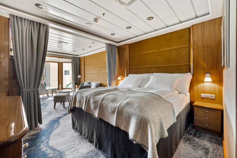 Hochwertig ausgestattete Grand Suite mit einem großen Doppelbett, weißen Bettdecken und grauen Decken. Der Raum ist mit Holzwänden und integrierten Nachttischen gestaltet, auf denen kleine Wandlampen für warmes Licht sorgen. Vorhänge trennen den Schlafbereich von einem angrenzenden Wohnbereich mit Sitzgelegenheiten, sichtbar im Hintergrund. Der Boden ist mit einem strukturierten Teppich versehen.