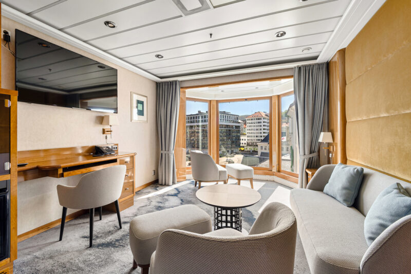 Wohnbereich einer Grand Suite mit einem großen Sofa, grauen Sesseln und einem runden Couchtisch. Große Panoramafenster mit Vorhängen bieten einen Ausblick auf die umliegende Landschaft mit Bergen und blauem Himmel. Auf der linken Seite befinden sich ein Schreibtisch und ein Spiegel. Die Suite ist modern eingerichtet, mit Teppichboden und warmen Holzelementen.