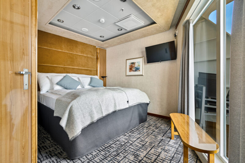 Modern eingerichtetes Schlafzimmer einer Expedition Suite mit einem großen Doppelbett, weißen Bettdecken und blauen Kissen. Holzelemente an Wänden und Möbeln verleihen dem Raum Wärme. Eine Glastür führt zu einem privaten Balkon, der einen Teil des Außenbereichs zeigt. An der Wand hängt ein Flachbildfernseher sowie ein dekoratives Bild. Der Boden ist mit einem gemusterten Teppich ausgestattet.