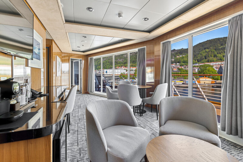 Heller, moderner Lounge-Bereich der Eigner Suite mit stilvollen grauen Sesseln, runden Holztischen und eleganten Dekorelementen. Große Panoramafenster bieten einen Ausblick auf einen grünen Hügel, ein Hafengebiet und blauen Himmel.