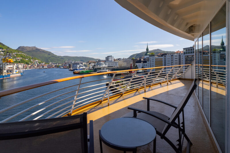 Privater Balkon der Eigner Suite mit zwei schwarzen Stühlen und einem kleinen Tisch. Der Balkon bietet einen beeindruckenden Blick auf einen sonnigen Hafen, umgeben von Hügeln, einer Stadtlandschaft und klarem blaue Wasser. Ein Geländer mit Glasabschnitten und die spiegelnde Balkontür verleihen dem Bereich ein modernes Design.