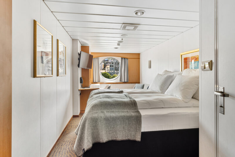 Helle Expedition Suite Q2 mit einem Doppelbett, das mit weißen Bettdecken und einer grauen Tagesdecke ausgestattet ist. Die Wände sind in Weiß gehalten und mit mehreren gerahmten Bildern dekoriert. Ein Flachbildfernseher ist seitlich angebracht, und ein Fenster am Ende des Raums bietet Blick auf die Umgebung.