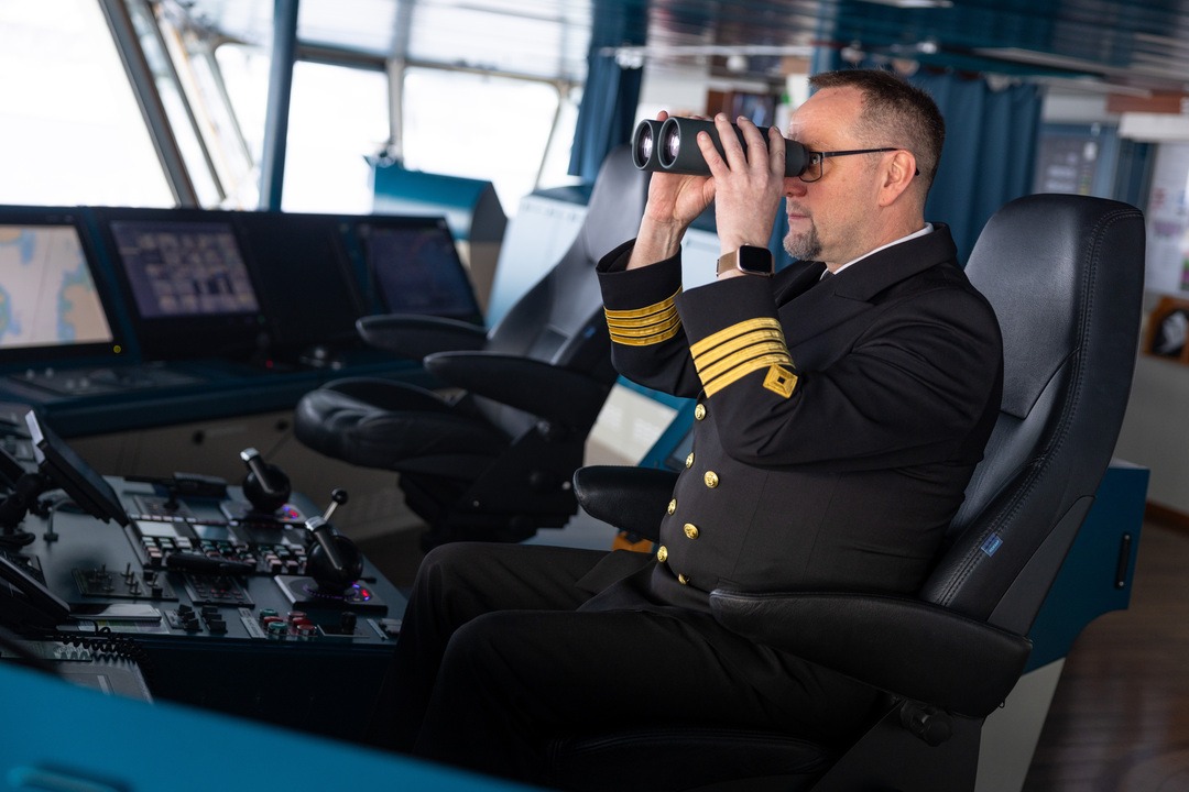 Ein Kapitän sitzt auf der Brücke eines Schiffes und schaut durch ein Fernglas. Er trägt eine marineblaue Uniform mit goldenen Streifen am Ärmel. Die Brücke ist mit verschiedenen Bildschirmen, Steuerungselementen und Instrumenten ausgestattet. Im Hintergrund sind große Fenster, durch die Tageslicht einfällt.