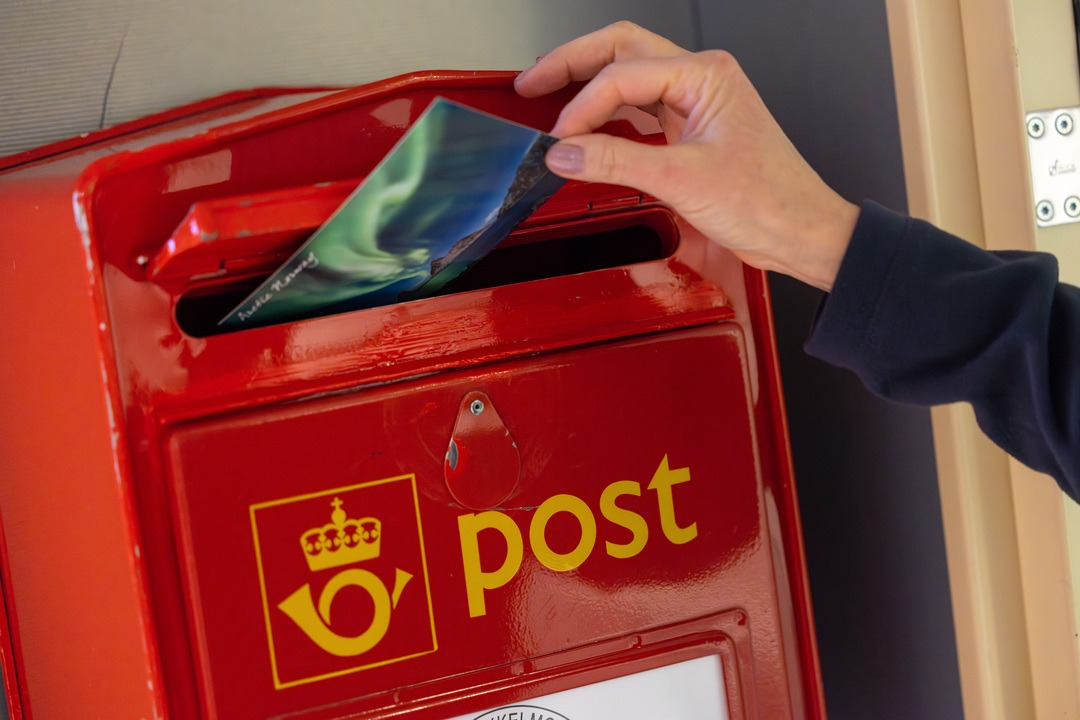 Eine Person steckt einen blauen Briefumschlag in einen roten Postbriefkasten. Der Briefkasten trägt das Logo und die Aufschrift „post“ in gelber Schrift. Der Hintergrund ist leicht verschwommen.