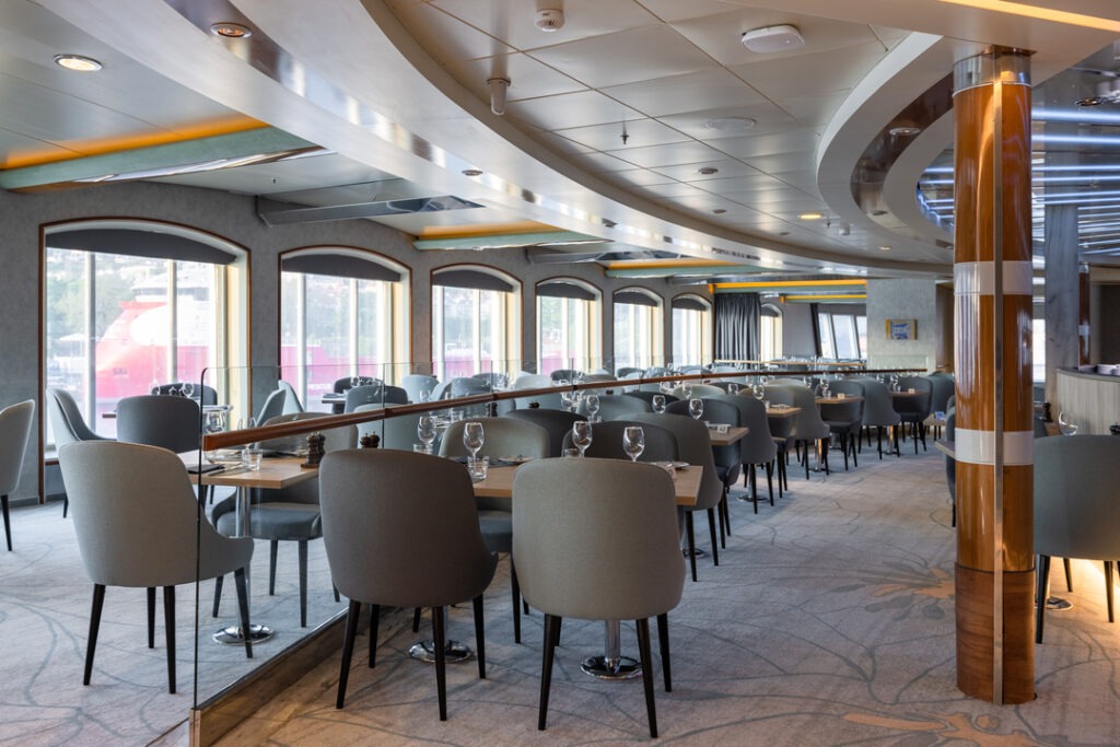 Ein modernes Restaurant mit mehreren Tischen und grauen Polsterstühlen. Große Fenster entlang der Wand lassen viel Tageslicht herein und bieten einen Blick nach draußen. Die Decke ist leicht gebogen, und Säulen mit goldenen Akzenten sind im Raum verteilt.