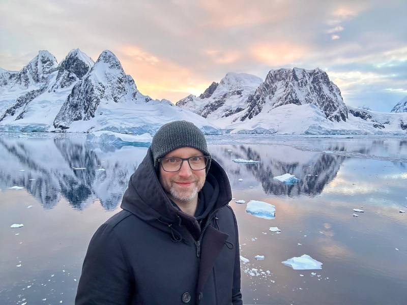 Timo von Eisrouten mit dunkler Jacke und Mütze steht vor einer eisbedeckten Landschaft mit schneebedeckten Bergen und schwimmenden Eisschollen in der Antarktis. Der Himmel zeigt sanfte Farbtöne, die sich im ruhigen Wasser spiegeln.