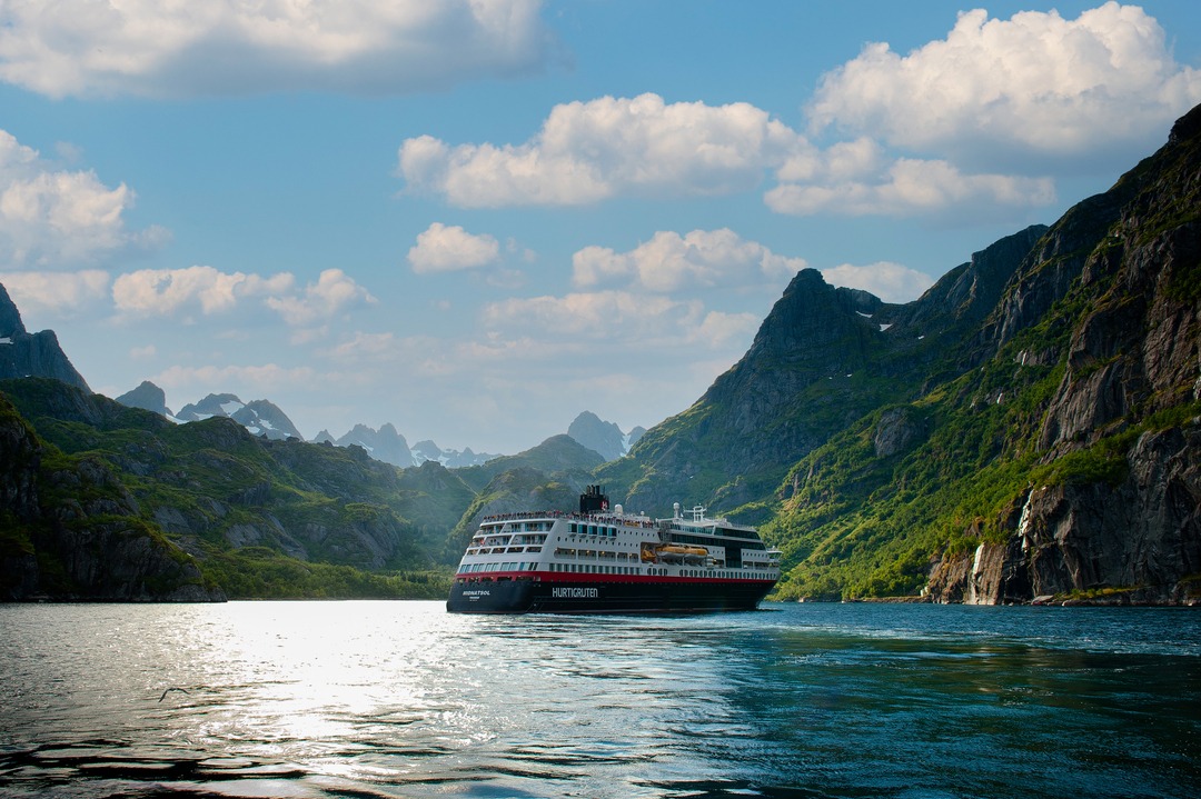 MS Midnatsol fährt durch einen sonnigen Fjord, umgeben von steilen, grünen Bergen und blauem Himmel mit weißen Wolken, typisch norwegische Naturkulisse.