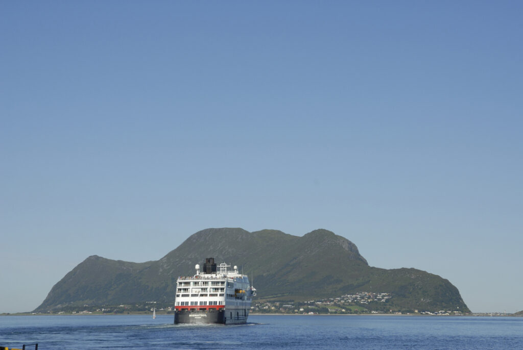 Die MS Midnatsol nähert sich einer grünen Insel unter klarem, blauem Himmel. Ruhige See und sommerliche Stimmung in der norwegischen Küstenlandschaft.