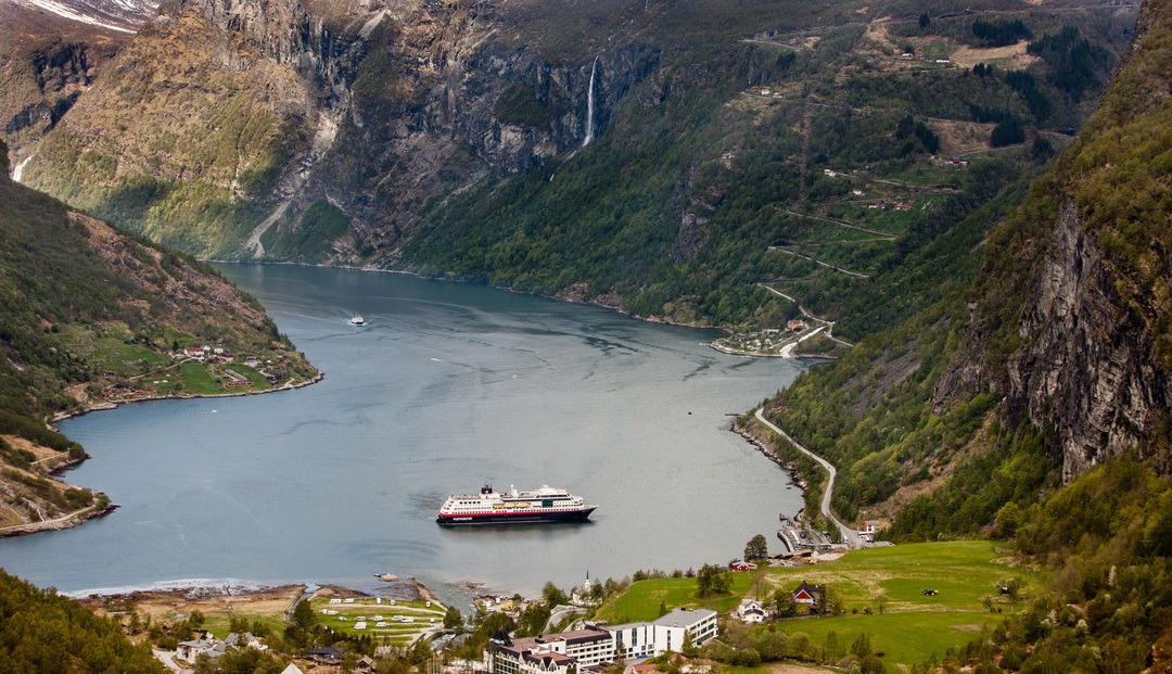 MS Midnatsol liegt im Geirangerfjord, umgeben von steilen Berghängen, Wasserfällen und grüner Landschaft. Ikonische Fjordlandschaft Norwegens von oben aufgenommen