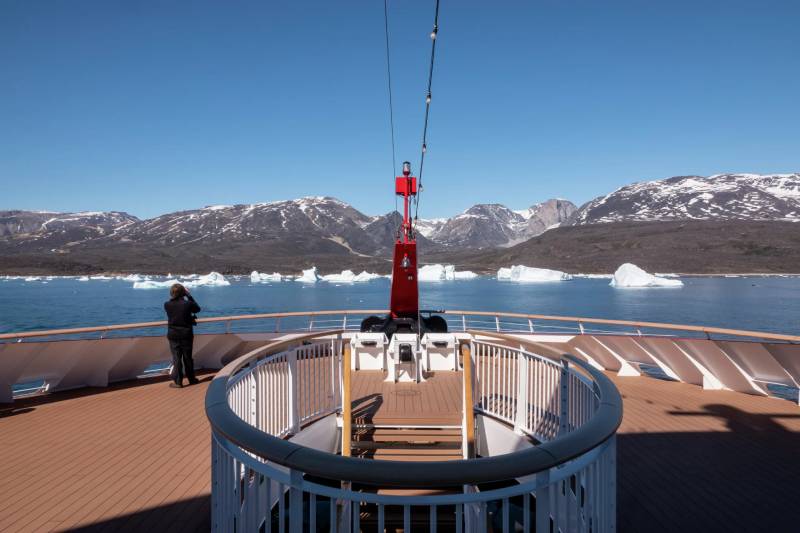 Blick vom Deck eines Expeditionsschiffs auf eine schneebedeckte Gebirgskette bei klarem Himmel; eine Person steht am Bug und genießt die Aussicht.