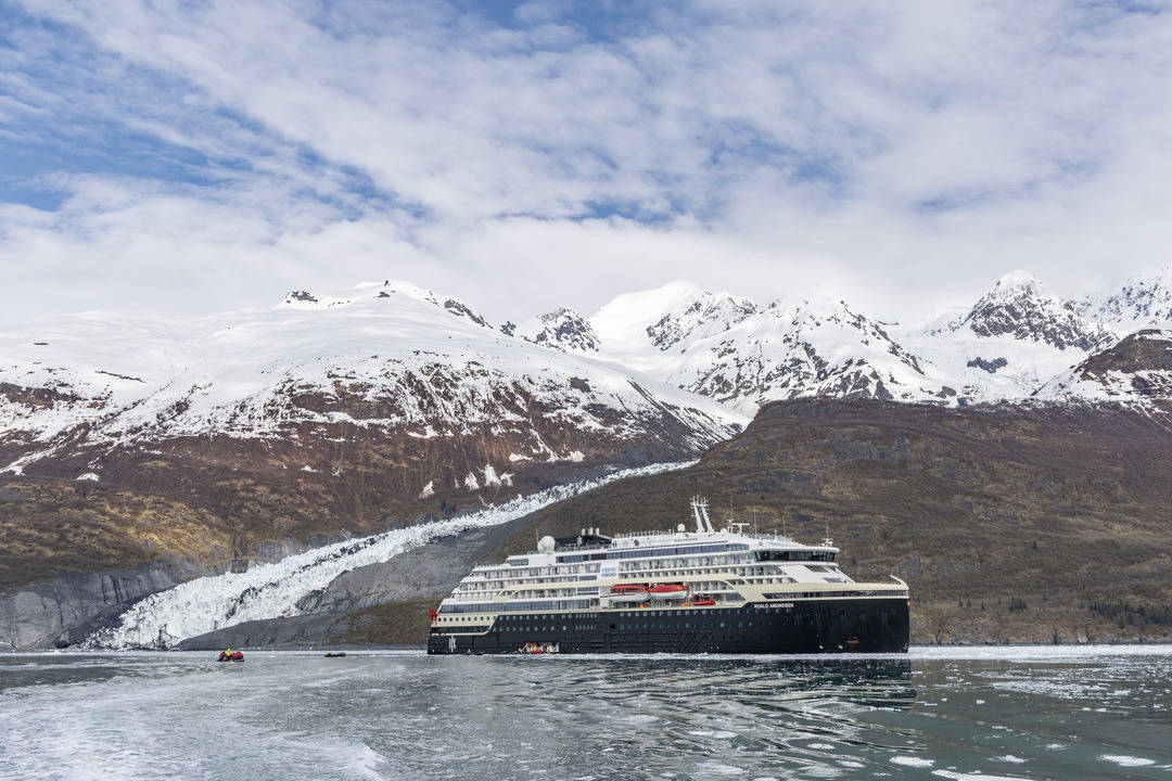 Das Expeditionsschiff MS Roald Amundsen fährt durch den College-Fjord in Alaska, umgeben von schneebedeckten Bergen und Gletschern unter teils bewölktem Himmel.