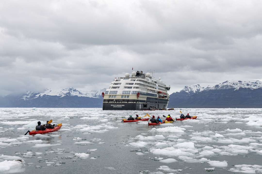 Mehrere Kajaks mit Passagieren paddeln zwischen Eisschollen, im Hintergrund liegt das Expeditionsschiff MS Roald Amundsen vor einer Bergkulisse unter bewölktem Himmel.