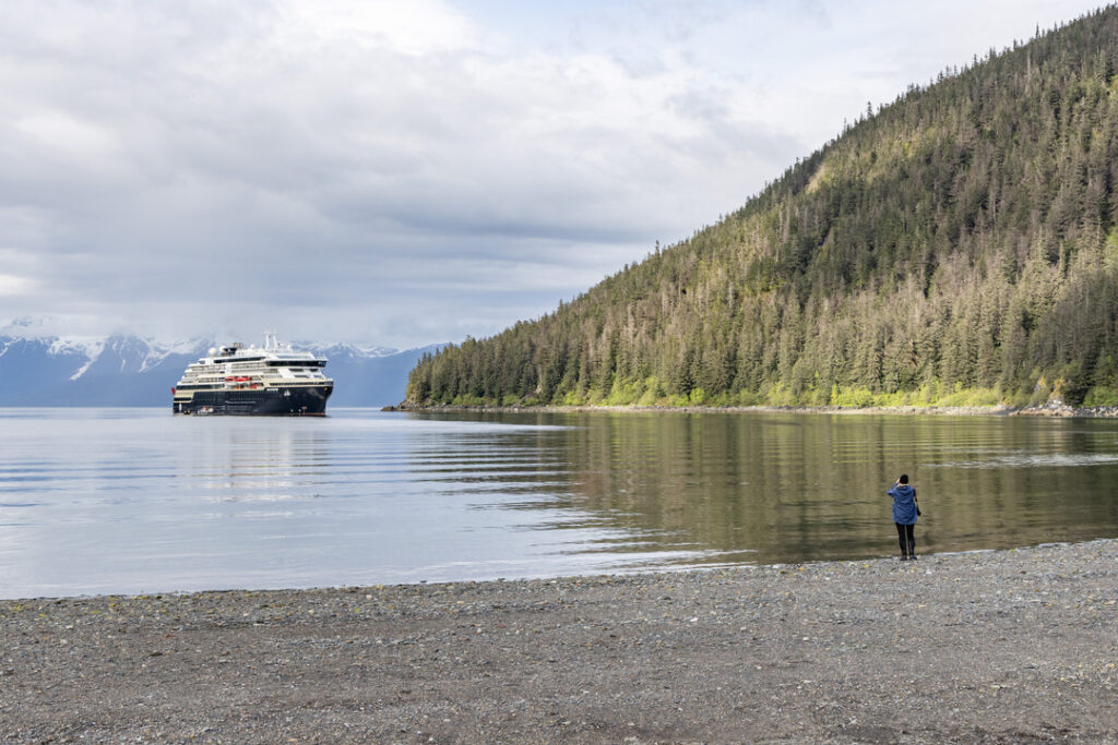 Eine Person steht am Kiesstrand in der William Henry Bay in Alaska, mit Blick auf die MS Roald Amundsen, die vor bewaldeter Küste vor Anker liegt.