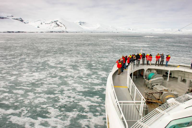 Eine Gruppe von Passagieren in roten HX-Jacken steht am Bug des Expeditionsschiffs MS Spitsbergen und beobachtet die eisbedeckte Landschaft in polarer Umgebung.