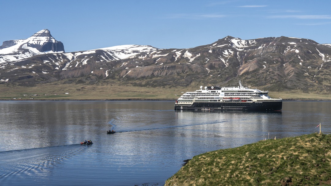 Die MS Fridtjof Nansen liegt vor Anker in der Bucht von Bakkagerði, Island, vor einer Kulisse aus grünen Hügeln und schneebedeckten Bergen.