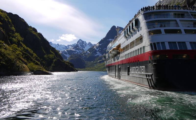 MS Midnatsol fährt durch einen norwegischen Fjord mit Bergen und Sonnenschein im Hintergrund.