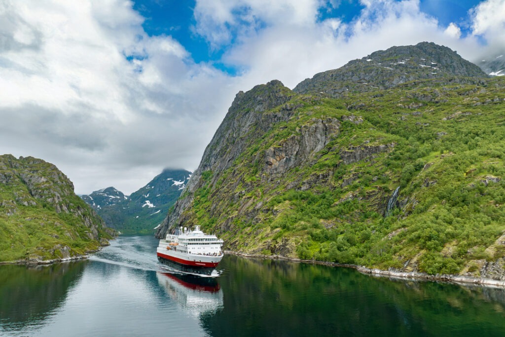 MS Finnmarken fährt durch einen engen Fjord, umgeben von hohen, grünen Bergen unter einem teils bewölkten Himmel.