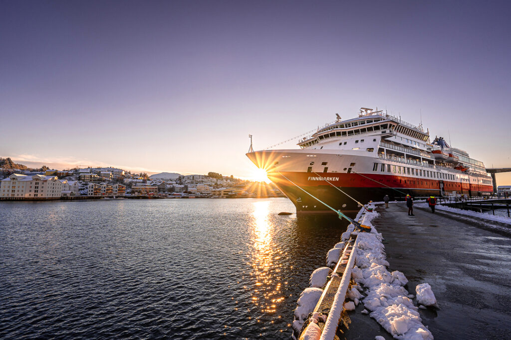 MS Finnmarken liegt bei Sonnenuntergang im winterlichen Hafen, umgeben von schneebedeckten Stegen und Gebäuden.