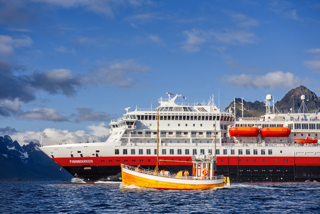 Die MS Finnmarken fährt bei klarem Wetter vor einer bergigen Küste, während ein kleines gelbes Boot im Vordergrund zu sehen ist.
