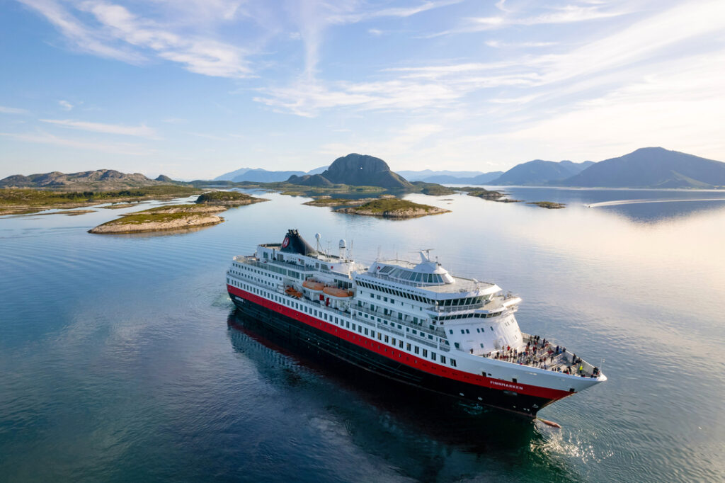 Hurtigruten Kreuzfahrtschiff MS Finnmarken fährt durch ruhige Gewässer, umgeben von Inseln und Bergen unter blauem Himmel; idyllische Landschaft bei Tageslicht.