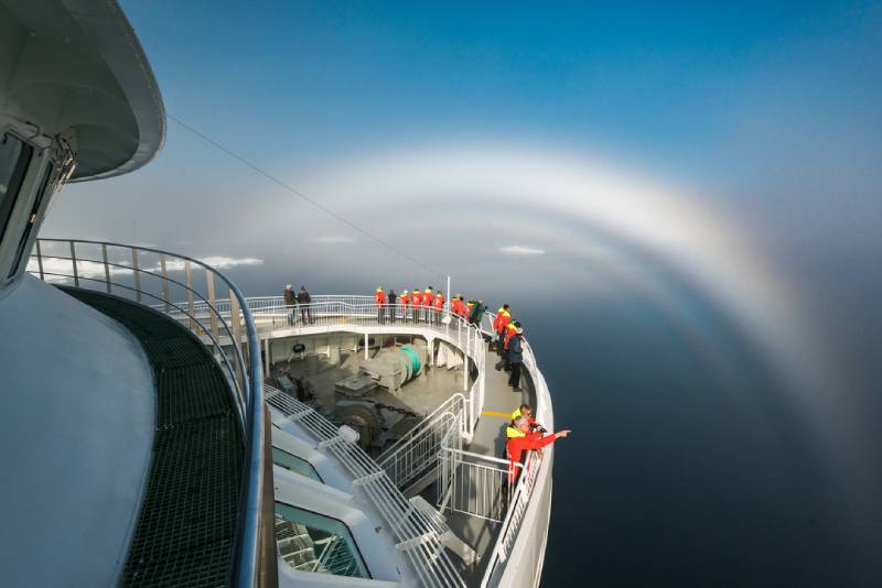 Passagiere in roten HX-Jacken stehen am Bug des Expeditionsschiffs MS Spitsbergen und beobachten einen seltenen Nebelbogen über dem ruhigen Meer.